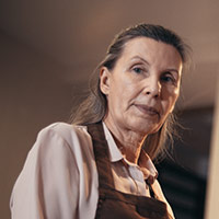 An image of a woman with grey hair and a brown apron and pink top on.